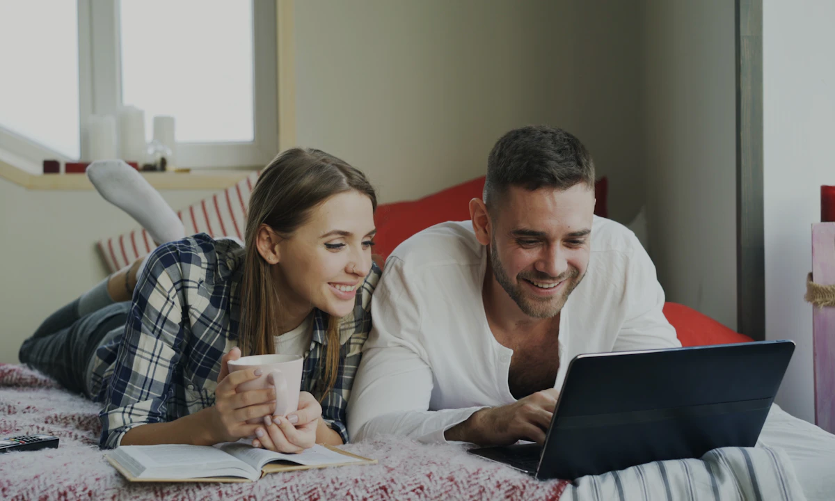 A smiling couple lying on a bed at home, looking at a tablet together